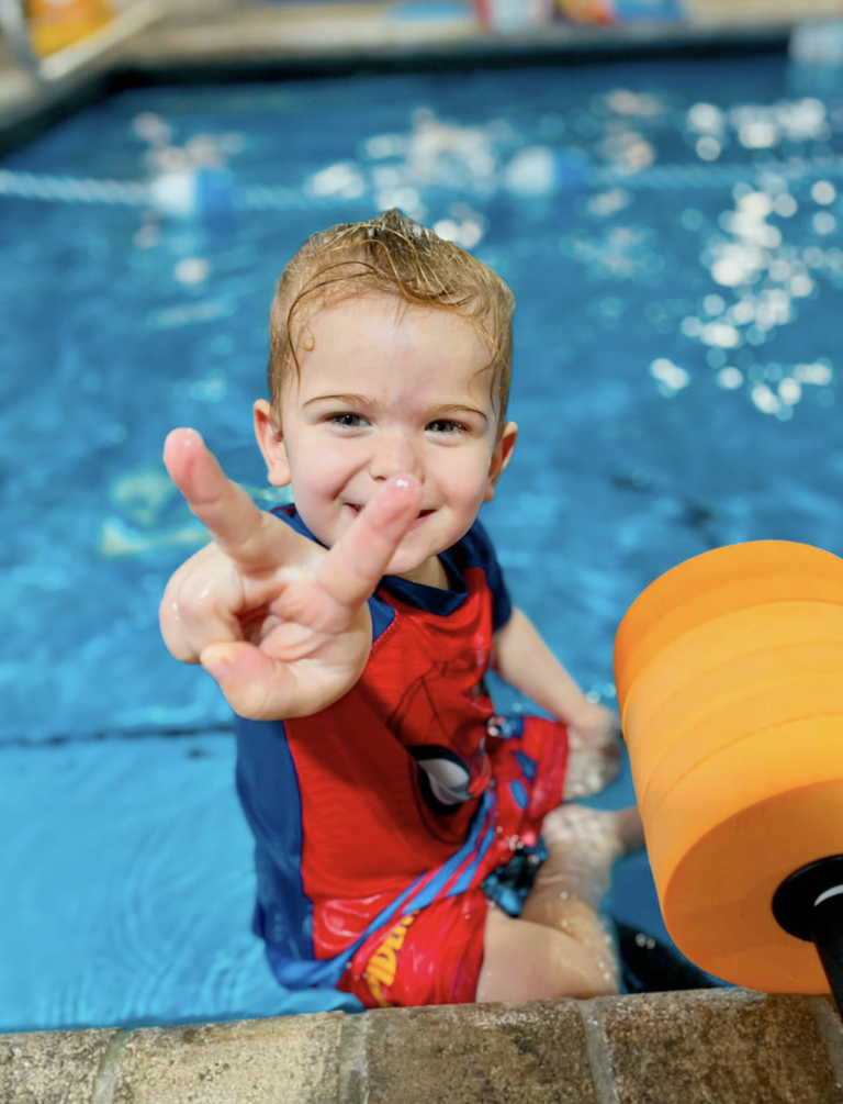 A young boy in the pool making a peace sign.